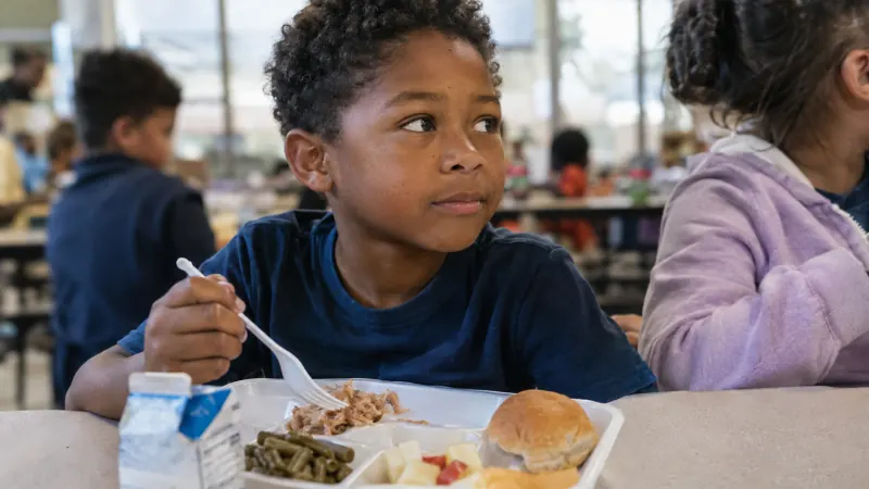 A school lunch tray with a small meal in front of a quiet child in a school cafeteria